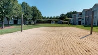 This community amenity space offers a well-rounded recreational and social environment designed to bring residents together. Outdoors, a full-size sand volleyball court provides a dedicated space for active play, set against a backdrop of lush green trees and open sky. Surrounding greenery and nearby structures create a pleasant, park-like atmosphere for outdoor gatherings and friendly competition.

The interior clubhouse is equally impressive, characterized by soaring vaulted ceilings adorned with striking exposed wooden beams that lend a warm, rustic-modern charm. Large multi-pane windows flood the space with abundant natural light, enhancing the bright, airy ambiance throughout. The main lounge area features a cozy fireplace, stylish tufted seating in rich jewel tones, and natural wood accent tables, creating an inviting setting for relaxation and socializing.

Adjacent to the lounge, a compact kitchen with dark cabinetry and stainless steel appliances supports events and gatherings with practical convenience. A dedicated business center offers workstation desks and ergonomic seating beneath the same dramatic vaulted ceiling, making it a functional yet refined space for residents to work or study. Hardwood-style flooring and wainscoting paneling tie the interior together, delivering a cohesive, polished aesthetic throughout.
