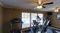This fitness space is designed to support an active lifestyle, featuring a thoughtfully arranged cardio zone with multiple exercise machines, including a treadmill, elliptical, and stationary bike. The room is bathed in natural light streaming through a row of large windows fitted with horizontal blinds, creating a bright and energizing atmosphere ideal for working out. A ceiling fan with an integrated light fixture is centrally mounted, ensuring consistent airflow and comfort during exercise sessions. Crown molding lines the upper walls, adding a refined architectural touch to the space. The textured ceiling and warm-toned walls contribute to an inviting yet purposeful ambiance. The flooring beneath the equipment provides a sturdy, practical surface suited for fitness use. Overall, this space balances functionality with comfort, offering a well-lit, ventilated, and organized environment for daily exercise routines.