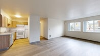 This single-floor residential unit offers a clean, functional layout designed for comfortable everyday living. The spacious living area is finished with light-toned wood-style flooring that extends throughout the common spaces, creating a bright and cohesive feel. A large multi-pane window floods the living room with natural light and provides views of the surrounding neighborhood, enhancing the open and airy ambiance of the space.

The galley-style kitchen is well-appointed with warm wood cabinetry offering generous upper and lower storage, a continuous countertop with an integrated sink, and a full suite of white appliances including a refrigerator, electric range, and dishwasher. The kitchen's light hardwood flooring complements the cabinetry tones for a warm, practical finish.

The bedrooms are carpeted for comfort and feature neutral wall tones that lend a calm, restful atmosphere. Each room is served by natural light through individual windows, and at least one bedroom includes a sliding-door closet with a wire shelving system for organized storage.

The entry area features durable wood-style flooring and convenient built-in closet storage adjacent to the front door. Throughout the unit, consistent gray baseboards and clean white walls give the space a modern, well-maintained character that is both practical and inviting.