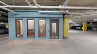 This large-scale indoor parking garage occupies a single level, offering an extensive field of clearly marked, numbered parking stalls across a smooth concrete floor. The structural design is defined by prominent yellow-painted concrete columns positioned throughout, providing both support and visual orientation across the open expanse. The exposed concrete ceiling reveals an organized network of pipes, conduits, and cable trays running overhead, alongside recessed and strip lighting that evenly illuminates the entire space.

A standout feature is a teal-painted enclosed core structure located within the garage. This unit houses what appears to be elevator access and utility rooms, with large glass-panel windows and gray metal doors providing interior visibility. The core is accessible from multiple sides and serves as a central service hub within the floor.

The garage includes dedicated accessible parking stalls marked with standard handicap signage, as well as electric vehicle charging stations integrated along the perimeter near the teal core. A gated vehicle entry and exit point with overhead clearance signage is visible, reinforcing the garage's functional, well-organized layout. The overall ambiance is clean, modern, and efficiently designed for high-capacity vehicle storage.