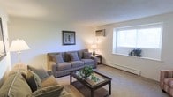 This single-floor space features a living room designed for comfort and practicality. The room is finished with wall-to-wall carpeting in a warm neutral tone, complementing the light-colored walls that create a bright and airy atmosphere. A window along one wall allows natural light to filter into the space, enhancing the open feel of the room. Climate control is provided by a wall-mounted air conditioning unit and a baseboard heating system, ensuring year-round comfort. The living area is well-proportioned, offering ample room for seating and a central coffee table with a glass top and dark wood frame, which serves as a functional focal point. Soft, neutral-toned upholstered seating adds to the relaxed ambiance. A framed piece of wall art and a table lamp with a warm glow contribute to the understated, welcoming character of the space. The overall design is straightforward and functional, with clean walls and simple finishes that provide a versatile foundation adaptable to a variety of furnishing styles and personal preferences.