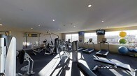 This fitness center is designed to accommodate a comprehensive workout experience within a single-level layout. The space is outfitted with a broad selection of exercise equipment, including multiple treadmills lined along the windowed wall, a multi-station cable and weight machine, and additional strength training apparatus positioned throughout. Dark rubberized flooring covers the workout area, providing both safety and durability underfoot. Recessed lighting in the white ceiling ensures consistent, even illumination across the room, complemented by natural light streaming through large windows that offer views of the surrounding landscape. Two flat-screen televisions are wall-mounted at comfortable viewing angles, allowing users to stay entertained during cardio sessions. A designated Training Zone area is visible along one wall, marked with signage and equipped with suspension training tools. The overall ambiance is functional and modern, with a clean, organized layout that maximizes usable floor space while keeping equipment accessible.
