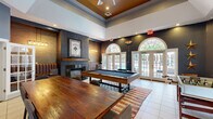 This well-appointed single-floor community clubhouse spans a generous footprint and offers a diverse range of amenities designed for both relaxation and activity. The centerpiece is a dramatic lounge area distinguished by soaring double-height ceilings with a warm, coffered wood accent, bold dark accent walls, and large arched windows flanked by French doors that flood the space with natural light and provide views to the outdoors. A classic brick fireplace anchors one wall, while a full-size billiards table and rustic wood communal table make this an ideal gathering spot.

Adjacent to the lounge, the kitchen and bar area features a long butcher-block countertop, stainless steel under-counter refrigerators, a draft tap, and a creative wall of reclaimed wood crate shelving — lending a distinctive industrial-rustic character. A separate living room nook offers a more intimate setting with bean bag seating and a wall-mounted TV, perfect for casual lounging.

The fitness center is expansive and well-lit, equipped with treadmills, stationary bikes, ellipticals, cable machines, free weights, and a weight bench. Full-length mirrors visually expand the space, and large windows overlook what appears to be a pool area, adding an energizing outdoor connection. Distressed wood-style flooring ties the fitness spaces together with a cohesive aesthetic throughout.