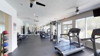 This fitness center offers a comprehensive workout environment designed to accommodate a variety of exercise needs. The space is laid out on a single floor with a generous open floor plan, finished with a dark, durable rubber flooring that provides both safety and easy maintenance. Large windows along one wall allow abundant natural light to fill the room while offering views of the surrounding outdoor greenery, creating an inviting atmosphere for exercise.

The facility is equipped with a well-rounded selection of cardio machines, including treadmills and elliptical trainers, arranged to maximize usable floor space. A multi-functional cable and weight machine anchors the strength training area, complemented by additional equipment visible throughout the room. Full-length mirrors line one wall, enhancing the sense of space while allowing users to monitor their form during workouts.

Overhead, multiple ceiling fans help maintain airflow and comfort throughout the facility. Recessed lighting and a textured ceiling complete the practical, clean aesthetic of the space. The overall ambiance is bright, functional, and well-maintained, making it a welcoming environment for residents seeking a convenient on-site fitness option.