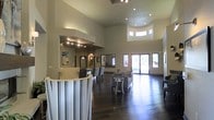 This single-story space makes a strong architectural impression through its dramatic, soaring ceilings that amplify the sense of openness throughout the interior. The main area is finished with rich, dark hardwood flooring that contrasts elegantly against the neutral, light-toned walls, creating a polished and welcoming atmosphere. Recessed lighting and pendant fixtures are thoughtfully positioned overhead to complement the natural light streaming in through large glass entry doors and a row of clerestory windows positioned high along the upper wall, further enhancing the airy, expansive feel of the space.

A curved reception or service counter anchors one side of the room, providing a functional focal point for visitors upon entry. Adjacent to it, a series of high-back dining or conference chairs are arranged along the counter, suggesting a multi-use purpose. The lounge area features upholstered seating arranged around a small side table, offering a comfortable setting for gatherings or informal meetings. Framed artwork along the walls adds a refined finishing touch. The large glass doors at the rear provide views of lush greenery outside, seamlessly connecting the interior to the outdoors.