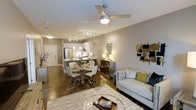 This single-floor residence showcases an open-concept design that integrates the living, dining, and kitchen spaces into one cohesive, flowing area. The living room is anchored by a contemporary sofa with accent pillows in neutral and yellow tones, complemented by a round glass coffee table and a decorative wall-mounted art piece, adding visual interest to the space. Warm wood-look flooring extends throughout, grounding the design with a sense of continuity.

The dining area features a round table paired with modern white chairs, positioned conveniently adjacent to the kitchen. The kitchen is well-appointed with stainless steel appliances, pendant lighting over the island, white cabinetry, and a track lighting system that ensures the workspace is well-lit. The island provides additional prep and seating space, making it a central hub for the home.

A ceiling fan with an integrated light fixture is centrally positioned overhead, offering both ventilation and ambient lighting. Neutral-toned walls throughout enhance the brightness of the space, while recessed lighting adds to the overall warm and welcoming ambiance of this thoughtfully designed residence.