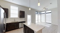 This single-story space showcases a thoughtfully designed open-concept layout where the kitchen and living areas flow seamlessly together. The kitchen is appointed with dark espresso cabinetry that contrasts elegantly against a crisp white quartz countertop on the central island, which serves as both a functional workspace and a natural gathering point. A classic white subway tile backsplash adds texture, while stainless steel appliances, including a dishwasher, contribute a polished, contemporary feel. Pendant lighting with cylindrical shades hangs above the island, adding focused illumination and a stylish accent.

The soaring ceilings amplify the sense of openness throughout, and a ceiling fan in the adjoining living area ensures comfort year-round. A striking bank of floor-to-ceiling windows with dark frames floods the living space with natural light, creating a bright, airy atmosphere. Additional windows near the kitchen sink and a white-trimmed interior door further enhance the light-filled character of the space. The neutral gray walls and light flooring tie the entire area together, resulting in a clean, modern, and inviting environment.