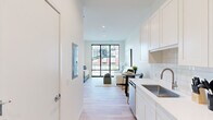 This thoughtfully designed single-floor residence showcases a clean, modern aesthetic throughout, with light hardwood flooring, crisp white walls, and recessed lighting creating a bright and airy atmosphere. The galley-style kitchen runs along one side of the main corridor and is outfitted with white shaker cabinetry extending to the ceiling, a white subway tile backsplash, a stainless steel sink, and a full suite of stainless steel appliances including a refrigerator, electric range, built-in microwave, and dishwasher. The kitchen flows seamlessly into the open living and dining area, which is anchored by a large floor-to-ceiling window that frames views of the surrounding urban streetscape and fills the space with abundant natural light. The bedroom is generously proportioned, featuring additional floor-to-ceiling windows and a narrow vertical window that allow natural light to flood the room from multiple angles, with continued views of the neighborhood. The bathroom is finished with large-format stone-look tile flooring, a sleek white vanity, and a spacious glass-enclosed walk-in shower with full-height tile surround. Every element of this residence reflects a cohesive, contemporary design sensibility with a focus on clean lines, quality finishes, and an open, light-filled environment.
