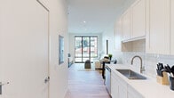 This thoughtfully designed single-floor residence showcases a clean, modern aesthetic throughout, with light hardwood flooring, crisp white walls, and recessed lighting creating a bright and airy atmosphere. The galley-style kitchen runs along one side of the main corridor and is outfitted with white shaker cabinetry extending to the ceiling, a white subway tile backsplash, a stainless steel sink, and a full suite of stainless steel appliances including a refrigerator, electric range, built-in microwave, and dishwasher. The kitchen flows seamlessly into the open living and dining area, which is anchored by a large floor-to-ceiling window that frames views of the surrounding urban streetscape and fills the space with abundant natural light. The bedroom is generously proportioned, featuring additional floor-to-ceiling windows and a narrow vertical window that allow natural light to flood the room from multiple angles, with continued views of the neighborhood. The bathroom is finished with large-format stone-look tile flooring, a sleek white vanity, and a spacious glass-enclosed walk-in shower with full-height tile surround. Every element of this residence reflects a cohesive, contemporary design sensibility with a focus on clean lines, quality finishes, and an open, light-filled environment.