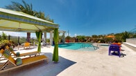 Located at 3510 N Craycroft Rd in Tucson, Arizona, this two-level outdoor amenity space offers a resort-inspired retreat set against the natural beauty of the Sonoran Desert. The centerpiece is a generously sized, freeform swimming pool with crystal-clear turquoise water, complemented by a separate spa and elegant water features including a pergola-mounted waterfall that creates a tranquil ambiance throughout the area. Expansive travertine pool decking surrounds the water features, providing ample space for lounge chairs and colorful outdoor seating arrangements. A shaded cabana with striped canopy and flowing green curtains offers a stylish retreat from the Arizona sun, while additional umbrella-shaded seating areas are distributed across the deck. Towering palm trees line the perimeter, reinforcing the resort atmosphere and providing natural shade. The upper level of the building features a covered outdoor terrace with views overlooking the pool area and the surrounding desert landscape. Textured retaining walls with a woven-block design add architectural interest to the perimeter, where EV charging stations and neatly landscaped desert plantings reflect a commitment to modern convenience and sustainability. The entire space is enclosed by decorative iron fencing, ensuring privacy while maintaining open views of the Tucson skyline.