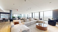 Located on an upper floor of a prominent high-rise at 1950 Broadway in Oakland, CA, this generously sized single-floor space delivers a striking first impression with floor-to-ceiling glass curtain walls that frame sweeping panoramic views of the Oakland cityscape, the San Francisco Bay, and the surrounding Bay Area. The open-plan layout is unified by warm-toned hardwood flooring and smooth white ceilings with recessed lighting, creating a bright and airy atmosphere throughout.

The space is organized into several distinct functional zones. A lounge area anchors one end of the floor, featuring an oversized sectional sofa, a circular wood coffee table, and sculptural accent seating — all bathed in natural light from the expansive glazing. A dramatic dark marble accent wall with integrated ambient lighting and a mounted flat-screen display serves as a bold focal point nearby.

The dining and gathering area accommodates multiple seating configurations, including a long formal dining table with upholstered chairs and a casual round table, complemented by a bar-height island with curved white seating. Cylindrical dark columns punctuate the open space, adding architectural rhythm without interrupting the flow. A built-in display niche with open shelving and a wet bar adds functional elegance. A welcoming entry foyer with lush greenery and velvet accent chairs completes this refined, versatile space.