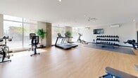 This fitness facility at 1819 Av Dollard in Montréal is a thoughtfully designed exercise space that provides a comprehensive workout environment. The room features light hardwood-style flooring throughout, smooth white walls, and recessed lighting that creates a clean, energizing atmosphere. Large floor-to-ceiling windows along one side of the gym flood the space with abundant natural light and offer open views of the surrounding outdoor greenery, enhancing the overall workout experience.

The cardio zone is well-appointed with multiple treadmills, a stationary bike, and an elliptical trainer, all positioned to take advantage of the expansive window views. For strength training, the gym offers a multi-function cable machine, a dedicated free weights area with a full rack of dumbbells ranging across multiple weight levels, and a flat adjustable bench. A large rubber mat defines the strength training zone, providing a safe and practical workout surface.

Additional amenities include exercise balls, yoga mats, and storage organizers for accessories, as well as a wall-mounted flat-screen television. A split-unit air conditioning system ensures a comfortable climate year-round. Exposed concrete columns add an industrial architectural touch that contrasts nicely with the otherwise bright and modern interior. A large mirror in the free weights area further enhances the sense of openness throughout this versatile fitness space.