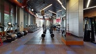 Located at 520 12th St S in Arlington, Virginia, this fitness center is a thoughtfully designed, single-floor facility that offers a comprehensive workout environment. The space is defined by soaring ceilings with an industrial-style dark grid panel finish, exposed red ductwork as a design accent, and linear LED lighting that illuminates the floor throughout. Large floor-to-ceiling windows wrap around key sections of the facility, allowing abundant natural light to flood in while offering views of the surrounding greenery.

The cardio zone is generously outfitted with treadmills, ellipticals, stair climbers, stationary bikes, and rowing machines, all arranged to take advantage of the natural light and outdoor views. The strength training area features multiple squat racks with barbells and weight plates, a full dumbbell rack, and a variety of cable and resistance machines positioned on durable rubber flooring. Floor-to-ceiling mirrors expand the sense of space throughout the strength area.

Sleek glass partition walls with black steel frames divide areas of the facility, including a separate lounge or studio space visible through the glass, featuring sculptural décor elements and a distinctive circular seating piece. Patterned carpet tiles in earth tones and blue define the transitional and cardio zones, while the overall design blends modern industrial aesthetics with functional, high-performance fitness amenities.