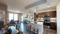 This single-floor residence showcases a thoughtfully designed open-concept floor plan that seamlessly connects the kitchen, dining, and living areas. The kitchen is a standout feature, equipped with rich dark-wood cabinetry, stainless steel appliances including a gas range and built-in microwave, and a sleek tile backsplash in a neutral palette. A generously sized white quartz island anchors the space, offering bar seating with upholstered stools and pendant lighting overhead, making it ideal for both meal preparation and casual dining.

The living area extends naturally from the kitchen, benefiting from abundant natural light streaming through large windows dressed with soft curtains. Wide-plank hardwood-style flooring runs throughout, adding warmth and visual continuity to the open layout.

A private balcony provides an elevated outdoor retreat, furnished with a bistro-style table and seating. The balcony offers sweeping views of the surrounding desert landscape, residential rooftops, lush tree canopies, and distant mountains beneath a vast, open sky — creating a serene and scenic outdoor environment perfect for relaxing or dining al fresco.