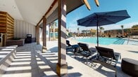 Located in Summerville, South Carolina, this single-level outdoor amenity space is designed to deliver a resort-style experience for its community. At the heart of the space is a large, sparkling swimming pool framed by an expansive paver deck finished in light-toned stone. Surrounding the pool, an array of lounge chairs and umbrellas provides ample space for relaxation under the South Carolina sun.

A covered colonnade runs along one side of the pool area, supported by substantial timber posts with metal base details and finished with a warm wood-paneled ceiling. This shaded corridor connects the main building to the pool deck, offering a comfortable transition between indoor and outdoor spaces. Large windows and floor-to-ceiling glass panels on the building façade allow for open sightlines and abundant natural light throughout.

An outdoor kitchen and lounge area is thoughtfully integrated into the design, featuring a mounted television, built-in grill, and a gated enclosure surrounded by slatted wood privacy screens and raised brick planters with lush greenery. The blend of natural wood, brick, and contemporary metal finishes gives the space a modern yet welcoming character. Palm trees and mature pines frame the perimeter, enhancing the relaxed, coastal atmosphere of this well-appointed community amenity.