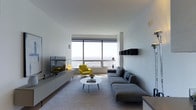 This single-floor space showcases a thoughtfully designed living area anchored by an expansive floor-to-ceiling window wall that frames striking panoramic views of the cityscape and open horizon beyond. The generous glazing floods the interior with abundant natural light, creating a bright and airy atmosphere throughout.

The living room is arranged around a large, low-profile gray sectional sofa that defines the seating area, complemented by a sleek oval white coffee table. A vibrant yellow accent chair introduces a bold pop of color against the otherwise neutral palette of whites, grays, and light wood tones. A wall-mounted flat-screen television sits above a low, minimalist media console along one wall, while a floating black shelf unit adds subtle visual interest on the opposite side.

The overall design ethos is decidedly minimalist, with clean lines, uncluttered surfaces, and a restrained color scheme that allows the dramatic exterior views to take center stage. Light-toned flooring and a soft area rug contribute to the cohesive, calm ambiance. The space feels open, well-proportioned, and refined, making it well-suited for both relaxed everyday living and stylish entertaining.