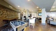 This single-floor community amenity space blends entertainment, work, and social gathering into one cohesive, well-designed environment. At the forefront of the room, a full-size foosball table anchors the game area, complemented by a shuffleboard table along the exposed brick accent wall — a striking design feature that adds an industrial, urban character to the space. Warm hardwood flooring flows throughout, tying together the various zones seamlessly.

A cluster of high-top tables with bar-style seating provides a casual dining and social area, while a dedicated workstation zone offers computer terminals and task seating for residents seeking a productive environment. Pendant lighting with Edison-style bulbs hangs above the central gathering areas, casting a warm, inviting glow that complements the recessed ceiling lighting throughout.

A wall-mounted flat-screen television is positioned to serve the lounge seating area, making it ideal for watching sports or entertainment. Framed artwork and a bold typographic mural on the brick wall add personality and a sense of place to the room. The combination of open layout, varied seating arrangements, and multi-purpose zones makes this an exceptionally versatile and welcoming communal space.