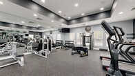 Located at 12167 Lincoln Lake Way in Fairfax, Virginia, this fitness center offers a thoroughly outfitted workout environment designed to support a wide range of exercise routines. The space features a contemporary aesthetic, with light gray walls, dark accent trim along the upper walls and ceiling, and durable speckled rubber flooring throughout — practical for heavy equipment and high-intensity use.

Floor-to-ceiling mirrors line multiple walls, enhancing the sense of space and allowing users to monitor their form. Recessed lighting provides bright, consistent illumination across the entire facility. Multiple windows fitted with slatted blinds allow natural light to filter in while maintaining privacy, offering views of the surrounding outdoor environment.

The cardio section is equipped with treadmills, elliptical machines, and a stair climber, all positioned to take advantage of the natural light. The strength training area includes a variety of selectorized weight machines, a full dumbbell rack with a wide range of weights, and a functional training station. For functional fitness and conditioning, the space also provides kettlebells, medicine balls, battle ropes, a large tire, TRX suspension trainers, and a dedicated wall-mounted storage rack. Wall-mounted televisions are positioned for convenient viewing during workouts, completing this well-rounded and thoughtfully organized fitness facility.