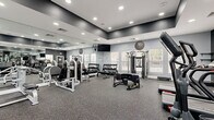 Located at 12167 Lincoln Lake Way in Fairfax, Virginia, this fitness center offers a thoroughly outfitted workout environment designed to support a wide range of exercise routines. The space features a contemporary aesthetic, with light gray walls, dark accent trim along the upper walls and ceiling, and durable speckled rubber flooring throughout — practical for heavy equipment and high-intensity use.

Floor-to-ceiling mirrors line multiple walls, enhancing the sense of space and allowing users to monitor their form. Recessed lighting provides bright, consistent illumination across the entire facility. Multiple windows fitted with slatted blinds allow natural light to filter in while maintaining privacy, offering views of the surrounding outdoor environment.

The cardio section is equipped with treadmills, elliptical machines, and a stair climber, all positioned to take advantage of the natural light. The strength training area includes a variety of selectorized weight machines, a full dumbbell rack with a wide range of weights, and a functional training station. For functional fitness and conditioning, the space also provides kettlebells, medicine balls, battle ropes, a large tire, TRX suspension trainers, and a dedicated wall-mounted storage rack. Wall-mounted televisions are positioned for convenient viewing during workouts, completing this well-rounded and thoughtfully organized fitness facility.