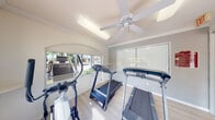 This single-room fitness center offers a clean, functional environment designed to support an active lifestyle. The space is finished with light wood-style flooring and crisp white walls accented by crown molding, lending a polished, well-maintained appearance throughout.

Natural light pours in through multiple large windows, which frame views of the surrounding outdoor amenities, including a pool area, palm trees, and landscaped grounds — creating an open, airy atmosphere that makes workouts feel less confined. A ceiling-mounted fan provides additional airflow and comfort during exercise sessions.

The room is equipped with several cardio machines, including multiple treadmills and an elliptical trainer, thoughtfully arranged to maximize the available floor space. A wall-mounted television is also present, offering entertainment during workouts.

Arched architectural details above the windows add a subtle decorative touch to the otherwise practical space. Vertical blinds on select windows allow for light and privacy control. An emergency exit sign and posted fitness center rules are visible near the entry door, which opens directly to the outdoor community area. Overall, this fitness center provides a bright, comfortable, and functional setting for daily exercise.