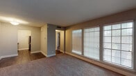 This single-floor unit offers a practical and functional layout with a cohesive neutral color palette throughout. The living area is generously proportioned, featuring wall-to-wall carpeting, large windows dressed with white horizontal blinds, and an accent wall clad in vertical shiplap-style paneling. Natural light filters in through multiple window panes, creating a bright and open atmosphere. A hallway with dark hardwood-style flooring connects the living space to the remaining rooms, providing a clear visual distinction between areas.

The galley kitchen is efficiently designed with white cabinetry lining both sides, light-speckled countertops, a mosaic tile backsplash, a stainless steel sink, and stainless steel appliances including a refrigerator and dishwasher. Dark hardwood-style flooring continues underfoot, complementing the clean, modern cabinetry.

The bedroom is a well-sized, carpeted room equipped with a ceiling fan with integrated lighting and a wide sliding-door closet featuring built-in shelving and hanging rods, offering ample storage.

The bathroom includes a white vanity with upper cabinetry, a wall-mounted mirror, tiled shower-tub combination, and continues the dark hardwood-style flooring seen throughout the non-carpeted areas.