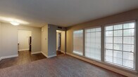 This single-floor unit offers a practical and functional layout with a cohesive neutral color palette throughout. The living area is generously proportioned, featuring wall-to-wall carpeting, large windows dressed with white horizontal blinds, and an accent wall clad in vertical shiplap-style paneling. Natural light filters in through multiple window panes, creating a bright and open atmosphere. A hallway with dark hardwood-style flooring connects the living space to the remaining rooms, providing a clear visual distinction between areas.

The galley kitchen is efficiently designed with white cabinetry lining both sides, light-speckled countertops, a mosaic tile backsplash, a stainless steel sink, and stainless steel appliances including a refrigerator and dishwasher. Dark hardwood-style flooring continues underfoot, complementing the clean, modern cabinetry.

The bedroom is a well-sized, carpeted room equipped with a ceiling fan with integrated lighting and a wide sliding-door closet featuring built-in shelving and hanging rods, offering ample storage.

The bathroom includes a white vanity with upper cabinetry, a wall-mounted mirror, tiled shower-tub combination, and continues the dark hardwood-style flooring seen throughout the non-carpeted areas.