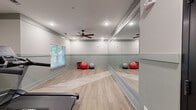 This dedicated fitness room is a thoughtfully designed workout space that offers a well-rounded environment for exercise. The room features light wood-style flooring laid in a herringbone pattern, which adds visual interest while remaining durable and easy to maintain. Two-tone walls in soft white and sage green are accented by a continuous chair rail, lending the space a clean, polished aesthetic.

A large floor-to-ceiling mirror spans one full wall, effectively doubling the perceived depth of the room and allowing users to monitor their form during workouts. Recessed lighting and a ceiling fan with dark wood-toned blades provide ample illumination and airflow, ensuring a comfortable environment during exercise. Natural light filters in through window blinds, adding brightness without excessive glare.

The space is equipped with multiple pieces of cardio equipment, including treadmills, a stationary bike, and a recumbent bike, offering variety for different fitness routines. Exercise balls are also present along the mirrored wall. A dark-framed barn-style door and a labeled mechanical closet are integrated into the layout, maintaining organization within the space. The overall ambiance is modern, clean, and purposefully designed to support an active and healthy lifestyle.