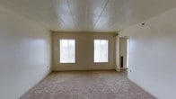This single-floor space presents a straightforward and functional layout centered around an open living area. The room is finished with wall-to-wall carpet in a neutral beige tone, providing a soft underfoot surface that complements the light-colored walls. A dropped panel ceiling adds a clean, uniform look overhead while housing recessed lighting that contributes to the overall brightness of the space.

Three windows line the main wall and an adjacent corner, allowing generous amounts of natural light to filter in and illuminate the interior. The windows are fitted with horizontal blinds, offering adjustable light control and privacy. The walls are painted in a warm off-white tone, creating a neutral backdrop adaptable to a variety of furnishing styles.

An open doorway on the right side of the main room leads to an adjoining space, suggesting additional rooms beyond the primary living area. A slim vertical element — likely a radiator or heating unit — is visible near the transition to the next room, indicating a dedicated heating system. The overall ambiance of the space is clean, understated, and move-in ready, offering a blank canvas for personalization.