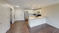 This single-floor unit is designed with a clean, modern aesthetic and a practical open layout. The kitchen serves as a central hub, featuring white shaker-style cabinetry, light granite-style countertops, stainless steel appliances including a refrigerator and range, and a large island with pendant lighting overhead and a built-in sink. Dark hardwood-style flooring flows throughout the main living areas, creating a warm contrast against the neutral beige walls.

The living areas are generously sized, with multiple windows allowing abundant natural light and views of the surrounding exterior. One room includes a sliding glass door that opens to a private balcony with outdoor views of greenery. The layout, as visible in the overhead floor plan, shows a well-organized arrangement of rooms connected by a central corridor.

Two bathrooms are present, each finished with large-format tile flooring, white vanity cabinetry, granite-style countertops, and walk-in showers with tile surrounds and handheld showerheads. One bathroom also includes a toilet and grab bar. A dedicated laundry room with side-by-side washer and dryer units and overhead shelving completes the space.