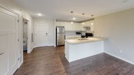 This single-floor unit is designed with a clean, modern aesthetic and a practical open layout. The kitchen serves as a central hub, featuring white shaker-style cabinetry, light granite-style countertops, stainless steel appliances including a refrigerator and range, and a large island with pendant lighting overhead and a built-in sink. Dark hardwood-style flooring flows throughout the main living areas, creating a warm contrast against the neutral beige walls.

The living areas are generously sized, with multiple windows allowing abundant natural light and views of the surrounding exterior. One room includes a sliding glass door that opens to a private balcony with outdoor views of greenery. The layout, as visible in the overhead floor plan, shows a well-organized arrangement of rooms connected by a central corridor.

Two bathrooms are present, each finished with large-format tile flooring, white vanity cabinetry, granite-style countertops, and walk-in showers with tile surrounds and handheld showerheads. One bathroom also includes a toilet and grab bar. A dedicated laundry room with side-by-side washer and dryer units and overhead shelving completes the space.