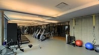 This modern fitness center is thoughtfully designed to accommodate a comprehensive workout experience. The main gym floor features durable, light-toned flooring and is lined with a robust selection of cardio machines, including rowing machines, elliptical trainers, and treadmills arranged in organized rows for ease of use. A dedicated strength training area runs along the perimeter, outfitted with a variety of weight machines and cable systems to support full-body resistance training. Toward one end of the space, a separate glass-partitioned studio area offers a warm hardwood floor, ideal for group classes, stretching, or personal training sessions. Functional training is also well represented, with TRX suspension straps anchored to the wall alongside a medicine ball and additional accessories. The gym is illuminated by recessed and linear overhead lighting that ensures a bright, energizing atmosphere throughout. Exposed concrete wall accents add an industrial character, while the clean, contemporary layout keeps the environment open and uncluttered. This facility is well-suited to support a wide range of fitness goals in a comfortable and professionally outfitted setting.