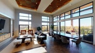 This single-floor space is defined by its striking architectural design, combining contemporary finishes with warm natural materials. Soaring ceilings are accented by a distinctive recessed slatted wood ceiling feature with integrated lighting, creating a refined focal point overhead. Dark-toned large-format tile flooring grounds the interior and flows cohesively throughout.

Floor-to-ceiling black-framed windows line the perimeter, flooding the space with abundant natural light and offering expansive views of the surrounding treetop and urban skyline. A lounge area anchors one side of the room, featuring a fireplace set within a built-in media niche flanked by warm wood cabinetry, complemented by leather accent chairs and a plush sofa. Adjacent to the lounge, a long dining table with upholstered chairs accommodates group gatherings beneath a modern chandelier.

A sleek wet bar with bar-height seating, pendant lighting, and a tiled backsplash provides a functional service area. Framed artwork adorns the walls nearby. Large sliding glass doors open directly to a furnished outdoor terrace with a wood-plank deck, covered seating areas, and panoramic views, seamlessly extending the interior living experience outdoors.
