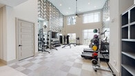 Located at 1401 Dixie Hwy in Covington, Kentucky, this fitness center is a thoughtfully designed, single-floor facility that combines contemporary aesthetics with full-featured workout amenities. The main exercise area commands attention with dramatically soaring double-height ceilings adorned with a bold geometric chevron-patterned accent wall, floor-to-ceiling mirrors, and recessed lighting complemented by a ceiling fan. A checkered tile floor in neutral tones runs throughout, adding visual interest while maintaining a clean, modern look. Large windows flood the space with natural light and provide views of the surrounding area, creating an open and energizing atmosphere. The strength training zone is well-appointed with a True Smith machine, a full rack of dumbbells, a weight bench, and a Bodycraft rowing machine, while a wall-mounted flat-screen TV offers entertainment during workouts. A separate area hosts dedicated resistance machines, including a leg extension machine, alongside built-in open shelving for storage. The cardio section features treadmills, an upright stationary bike, and additional cable machines, positioned near glass doors and windows for ample daylight. The facility connects to a corridor leading to additional amenity spaces, rounding out this polished and comprehensive fitness destination.