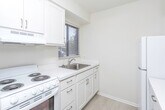This single-floor unit in Royal Oak, Michigan, offers a well-organized layout designed for comfortable everyday living. The kitchen is efficiently arranged with white cabinetry, white laminate countertops, and a stainless steel sink positioned beneath a window fitted with horizontal blinds, allowing natural light to brighten the workspace. A white electric coil range with an overhead exhaust hood and a full-size refrigerator complete the kitchen's appliances, providing all essential cooking amenities.

The living area features neutral-toned carpet flooring and is illuminated by a central pendant light fixture. A pass-through opening connects this space to the kitchen, enhancing the sense of openness. A sliding glass door leads directly to an outdoor green area, bringing in abundant natural light and providing easy exterior access.

The bedroom is carpeted and features a window that allows natural light to enter, keeping the space bright and airy.

The bathroom is finished with small-format mosaic tile flooring and includes a sliding glass-door shower enclosure with a grab bar, a toilet, and a white vanity with an integrated sink and a large mirror above. The clean, neutral palette throughout the unit creates a cohesive and welcoming atmosphere.