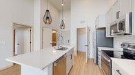 This single-floor unit showcases a contemporary design defined by soaring double-height ceilings and an open-concept layout that maximizes the sense of space and airflow. Light wood-tone flooring runs throughout, complementing the crisp white walls and black accent hardware found on doors and fixtures.

The kitchen serves as the central feature of the living area, anchored by a generously sized white quartz island with an undermount sink and a commercial-style spring faucet. White subway tile backsplash, gray shaker-style cabinetry, and a full suite of stainless steel appliances—including a range, microwave, refrigerator, and dishwasher—complete the space. Two geometric black cage pendant lights hang above the island, adding a modern industrial touch.

The living area benefits from an impressive bank of floor-to-ceiling windows that flood the space with natural light and offer elevated city views. A dedicated in-unit laundry area houses a stacked washer and dryer. The bathroom features a white vanity with matte black hardware and direct access to a spacious walk-in closet with adjustable shelving and hanging rods. Additional bedrooms are bright, well-proportioned, and fitted with black-framed windows.