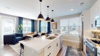 This single-floor residence showcases a thoughtfully designed open-concept layout with contemporary finishes throughout. The kitchen serves as a central hub, anchored by a large white quartz island with an undermount sink and seating for multiple bar-height chairs. Three matte black pendant lights hang above, complementing the white shaker cabinetry, patterned tile backsplash, stainless steel appliances, and a sliding barn door that adds a modern-rustic touch. Built-in open shelving flanks the kitchen area, providing both storage and display space.

The living area flows seamlessly from the kitchen and is bathed in natural light through oversized windows framed by navy curtains, revealing expansive city skyline views. A glass door provides direct access to an outdoor balcony, enhancing the connection to the urban surroundings.

The bedroom continues the light-filled theme with a large window and balcony door, offering additional city views. Two full bathrooms serve the residence — one features dark slate-style tile flooring with a white vanity, while the primary bath includes a dual-sink vanity with matte black fixtures and a large framed mirror. Wood-look flooring runs throughout the main living spaces, tying the design together cohesively.