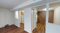 This single-floor residence offers a well-organized and inviting layout designed for comfortable everyday living. The interior is finished with rich hardwood-style flooring that runs throughout, adding warmth and continuity across the space. One of the most distinctive architectural features is a full exposed brick accent wall, which lends character and a classic, timeless aesthetic to the main living area.

The open layout allows for a natural flow between the living area and the kitchen, which is visible through a wide archway. The kitchen is equipped with wooden cabinetry and includes a full-size refrigerator, as well as a window that allows natural light to brighten the space. A set of bifold closet doors provides convenient built-in storage, keeping the living area neat and functional.

White-painted walls and trim contrast cleanly against the warm tones of the brick and flooring, creating a balanced and cohesive interior palette. The textured ceiling adds a subtle traditional detail throughout. Ceiling-mounted HVAC vents indicate a central air system for year-round comfort. Overall, the space combines practical functionality with characterful design elements, making it a well-rounded and visually appealing single-floor residence.
