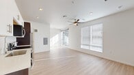 This efficiently designed single-floor unit offers a clean, contemporary aesthetic throughout its thoughtfully arranged layout. The open-concept kitchen and living area serves as the heart of the space, bathed in natural light streaming through large windows fitted with white horizontal blinds. A ceiling fan overhead provides both comfort and a subtle design accent. Light-toned hardwood flooring runs seamlessly throughout, lending warmth and a sense of continuity to the interior.

The kitchen is well-appointed with stainless steel appliances, including a refrigerator, electric range, built-in microwave, and dishwasher. Two-toned cabinetry — white upper cabinets paired with dark lower cabinets — offers ample storage, while light countertops provide a bright, functional workspace. Recessed lighting illuminates the kitchen area effectively.

The bathroom continues the modern design language with a dark wood vanity, white countertop, undermount sink, brushed nickel fixtures, and a round mirror that adds a contemporary touch. Light hardwood flooring carries into this space as well.

The living area is open and versatile, with multiple windows ensuring strong natural light and a connection to the outdoors. A dedicated closet provides practical storage. The overall ambiance is bright, minimal, and move-in ready.