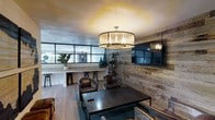 This single-floor space combines rustic and industrial design elements to create a sophisticated and inviting lounge environment. The most striking architectural feature is the reclaimed wood accent wall, which spans a significant portion of the room and provides a warm, textured backdrop for the wall-mounted flat-screen television. This natural wood element contrasts beautifully with the smoother finishes found throughout the rest of the space.

The seating area is anchored by a dark metal coffee table surrounded by rich brown leather club chairs, lending the space a classic yet contemporary feel. Overhead, a large drum-style pendant light with a fabric shade and interior candelabra bulbs serves as a central focal point, casting warm ambient light across the room.

Adjacent to the main lounge area, a bar-height counter with backless stools runs along a glass partition wall, allowing natural light to filter in from the neighboring space while maintaining a degree of visual separation. Light hardwood flooring ties the zones together seamlessly.

Decorative touches, including tall vases and potted greenery, add organic warmth to the overall industrial-chic aesthetic of the space.