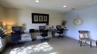 This single-floor business center offers a practical and comfortable environment designed to support productivity. The room features a long, dark-surfaced workstation desk that runs along the wall, accommodating multiple computer setups complete with monitors, keyboards, and printers — providing ample resources for a variety of tasks. Ergonomic black office chairs are positioned at each workstation, ensuring comfort during extended use. The space is finished with neutral-toned walls and textured carpeting that adds warmth underfoot. Recessed ceiling lights provide consistent, even illumination throughout the room, complemented by natural light that filters in and casts a soft pattern across the floor. A framed decorative triptych artwork anchors the main wall, adding a touch of visual interest to the otherwise functional setting. Decorative accents such as small floral arrangements and figurines are thoughtfully placed along the desk, lending a welcoming atmosphere. A large wall clock is mounted nearby, keeping occupants mindful of time. The overall ambiance is clean, organized, and professional — making this business center a convenient and well-appointed resource for anyone in need of a dedicated workspace.
