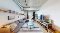 This two-bedroom, two-bathroom residence at The Smyth showcases a distinctive industrial-modern aesthetic throughout its well-appointed single-floor layout. Exposed concrete ceilings and visible ductwork define the architectural character of the space, complemented by light wood flooring that runs throughout the main living areas.

The open-concept living room offers generous proportions, featuring large floor-to-ceiling windows and a sliding glass door that invite abundant natural light and provide views of the surrounding cityscape. The kitchen is outfitted with gray cabinetry, white quartz countertops, a white subway tile backsplash, and a full suite of stainless steel appliances, including a gas range. A breakfast bar with seating for three connects the kitchen to the living area.

The primary bedroom continues the industrial aesthetic with exposed concrete ceilings, large windows, and ample space. The second bedroom is equally well-sized and shares the same design sensibility. Both bathrooms are finished with large-format tile flooring and modern fixtures. The primary bath features a sleek vanity with a bold framed mirror, while the second bath includes a soaking tub with white tile surround and matte black hardware throughout.