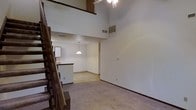 This two-story space is defined by its dramatic double-height ceiling in the main living area, which creates a strong sense of openness and volume. A wood-banistered staircase runs along one side of the room, leading to the upper level, where an open loft-style overlook peers down into the living space below. The ceiling fan on the upper level and the HVAC vent positioned high on the wall underscore the impressive vertical scale of the room.

The main living area is finished with wall-to-wall carpeting in a neutral tone, providing a soft, understated foundation throughout. A peninsula-style breakfast bar separates the living space from the kitchen area, which is visible in the background and illuminated by a pendant light fixture. The kitchen features dark countertops that contrast with the lighter cabinetry, and a passthrough design keeps the layout connected and functional.

The overall ambiance is clean and minimal, with white walls that reflect light throughout the interior. The open sightlines between floors and the interplay between the lower and upper levels give the space a versatile, multi-dimensional character well-suited to a variety of uses.