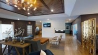 This well-appointed single-floor space showcases a thoughtfully designed interior that blends modern aesthetics with comfortable functionality. The centerpiece of the main lounge area is a sleek, built-in linear fireplace set into a crisp white wall, flanked by a mounted flat-screen TV — creating an inviting focal point for gathering and relaxation. Seating is arranged with intention, featuring a mix of plush sofas, tufted leather ottomans, and minimalist accent chairs atop a neutral area rug, encouraging both casual conversation and quiet retreat. The ceiling draws immediate attention with its distinctive wood-paneled tray design, complemented by an eye-catching sputnik-style chandelier and recessed lighting that together create warm, layered illumination throughout the space. Rich hardwood flooring extends across the entirety of the room, unifying the open layout. Bold teal and charcoal accent walls add depth and visual interest, while framed artwork contributes a curated, gallery-like quality. Adjacent to the main lounge, a kitchenette with stainless steel appliances and cabinetry provides practical convenience. A connecting corridor with continued accent wall detailing leads to additional areas, reinforcing the space's cohesive and polished design language.