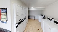 This single-floor shared laundry facility in Abilene offers a functional and well-equipped space designed to serve multiple users efficiently. The interior features light-toned tile flooring throughout, contributing to a clean and easy-to-maintain environment. The main laundry area is outfitted with multiple stacked dryer units along one wall and a row of top-load washing machines, providing ample capacity for high-volume use. A built-in folding counter offers a convenient surface for sorting and handling laundry. The space is finished with white walls and ceiling, keeping the atmosphere bright and neutral. A large picture window near the entrance allows natural light to filter in and provides an open view of the tree-lined sidewalk and neighboring structures outside. The entry area includes multiple door access points and features a mounted fire extinguisher, reflecting attention to safety compliance. Overhead lighting ensures the space remains well-lit at all hours. The layout is straightforward and accessible, with clear circulation paths between the machines and folding area, making the facility practical and user-friendly for daily use.