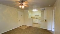 This single-story space is designed with a functional, open-concept layout that connects the living area directly to the kitchen. The living room features wall-to-wall carpet flooring and is centered around a ceiling fan with an integrated light fixture, providing both comfort and illumination. A front entry door opens directly into this main living space, which offers a clean, neutral palette with beige walls and white trim throughout.

The kitchen is positioned at the rear of the living area and is separated by a half-wall partition that also serves as a breakfast bar with a tiled countertop overhang. The kitchen itself is outfitted with white cabinetry, white appliances including a dishwasher and refrigerator, a single-basin sink, and an overhead range ventilation unit. The white cabinetry and countertops contribute to a bright, cohesive aesthetic.

A separate room — likely a bedroom — continues the carpet flooring and neutral wall tones. It includes a ceiling fan with a light kit and a window dressed with horizontal blinds, allowing filtered natural light and outdoor views. The overall ambiance of the space is straightforward, clean, and move-in ready, with consistent finishes throughout.