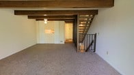 Located at 5312 Gatehouse Dr in Columbus, Ohio, this two-story home offers a distinctive architectural character defined by its prominent exposed wood beam ceilings, which add warmth and a rustic charm to the interior. The main living area is carpeted and open in layout, providing a comfortable and functional gathering space. A striking open-tread wooden staircase with a black metal safety gate serves as both a practical and visual focal point, connecting the two floors in an airy, transparent fashion. Adjacent to the living space, a sliding panel partition leads toward the kitchen area, where a pass-through window can be seen, enhancing the flow between spaces. A ceiling fan adds to the practical comfort of the main level. The entryway is visible from the living area, featuring a classic white door that welcomes natural light into the corridor. The consistent use of wood tones throughout — from the ceiling beams to the staircase framing — creates a cohesive, earthy aesthetic across the home. The overall ambiance is straightforward and functional, with architectural details that give the space character and a sense of established, enduring design.
