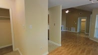 This single-floor residence offers a thoughtfully designed open layout that promotes a natural flow between living spaces. The interior is finished in warm, neutral tones throughout, creating a welcoming and cohesive ambiance. Hardwood-style flooring extends across the main living area, adding warmth and visual continuity, while tile flooring defines the kitchen zone, providing a practical transition between spaces. The living area features vaulted ceilings that lend a sense of openness and volume, complemented by a ceiling fan that enhances both comfort and style. A glimpse into the kitchen reveals wood-toned cabinetry, offering ample storage in a classic, functional design. Natural light filters in through a window, brightening the interior and highlighting the clean lines of the space. Neutral wall colors and white trim work together to create a fresh, timeless aesthetic. Floor vents are thoughtfully integrated throughout, ensuring efficient climate control. The overall ambiance is calm and understated, providing a solid foundation for a variety of interior styles and furnishing preferences.