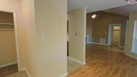 This single-floor residence offers a thoughtfully designed open layout that promotes a natural flow between living spaces. The interior is finished in warm, neutral tones throughout, creating a welcoming and cohesive ambiance. Hardwood-style flooring extends across the main living area, adding warmth and visual continuity, while tile flooring defines the kitchen zone, providing a practical transition between spaces. The living area features vaulted ceilings that lend a sense of openness and volume, complemented by a ceiling fan that enhances both comfort and style. A glimpse into the kitchen reveals wood-toned cabinetry, offering ample storage in a classic, functional design. Natural light filters in through a window, brightening the interior and highlighting the clean lines of the space. Neutral wall colors and white trim work together to create a fresh, timeless aesthetic. Floor vents are thoughtfully integrated throughout, ensuring efficient climate control. The overall ambiance is calm and understated, providing a solid foundation for a variety of interior styles and furnishing preferences.