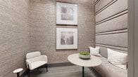 This refined single-floor space is thoughtfully designed to maximize both function and aesthetic appeal within its intimate footprint. The dining area serves as the centerpiece, featuring a sleek round pedestal table with a deep navy base, surrounded by plush cream upholstered chairs. A built-in banquette with a tall, channel-tufted headboard anchors one side of the room, accented by coordinating throw pillows and a decorative moss bowl centerpiece. Textured grasscloth-style wall coverings in warm taupe tones lend depth and sophistication throughout, while a pair of framed abstract artworks stacked vertically adds a gallery-quality finishing touch. Natural light enters generously through a full-height sliding glass door fitted with floor-to-ceiling drapery panels, offering views to an exterior courtyard. A separate lounge chair provides additional seating flexibility. The corridor connecting the dining area to the bathroom showcases herringbone-patterned hardwood flooring, recessed cove lighting with warm accents, and framed wall art in dark wood frames. The bathroom is accessed through glass-paneled dark-framed doors, maintaining visual continuity with the space's polished, contemporary design language throughout.