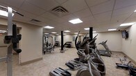 The Basketball Court & Fitness Center offers a comprehensive recreational facility designed to support a wide range of workout routines. The fitness area is housed within a spacious, open-plan room featuring a drop ceiling with recessed fluorescent panel lighting that evenly illuminates the entire space. Neutral-toned walls and a light-colored tile floor contribute to a clean, functional aesthetic throughout.

The gym is equipped with a robust selection of exercise equipment, including a row of elliptical trainers and treadmills along one side, providing ample cardio options. A multi-station strength training machine anchors one end of the room, offering a variety of resistance-based exercises. A free weights section features a well-organized dumbbell rack stocked with multiple weight increments, alongside additional benches and functional training equipment. Structural columns are positioned throughout the floor, adding an architectural element while maintaining open sightlines across the room. The layout ensures sufficient space between equipment stations, allowing for comfortable movement and simultaneous use by multiple individuals.